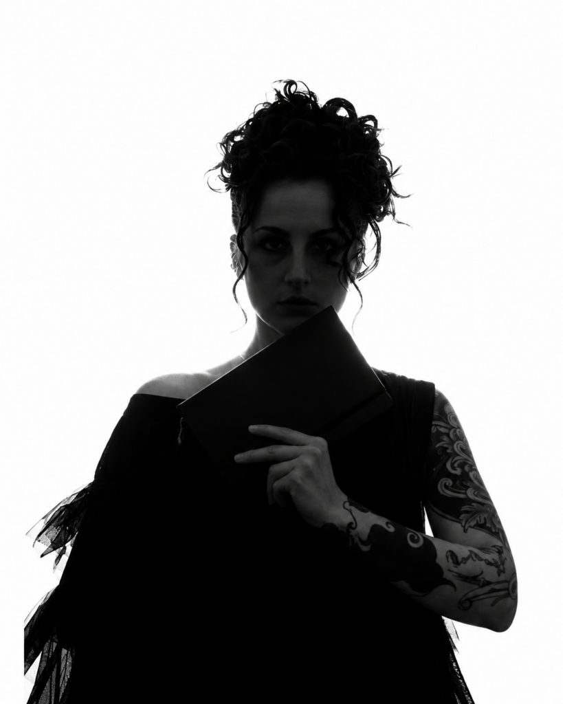 Anna Caminiti in bianco e nero, con capelli raccolti e un portfolio nero tra le mani, su sfondo bianco.